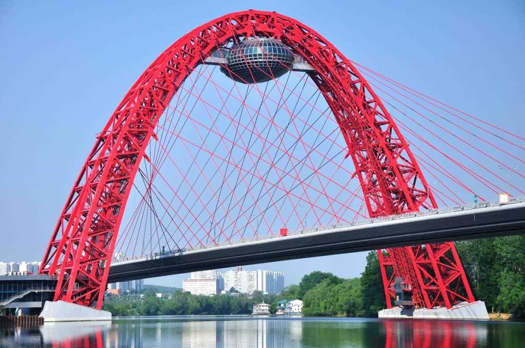 Живописный мост – яркое украшение столицы России и одно из самых концептуально примечательных инженерных сооружений в мире Мост расположен на стыке Москвы-реки и двух рукотворных водных спрямлений, выделяясь на фоне городского пейзажа идеальной алой параб